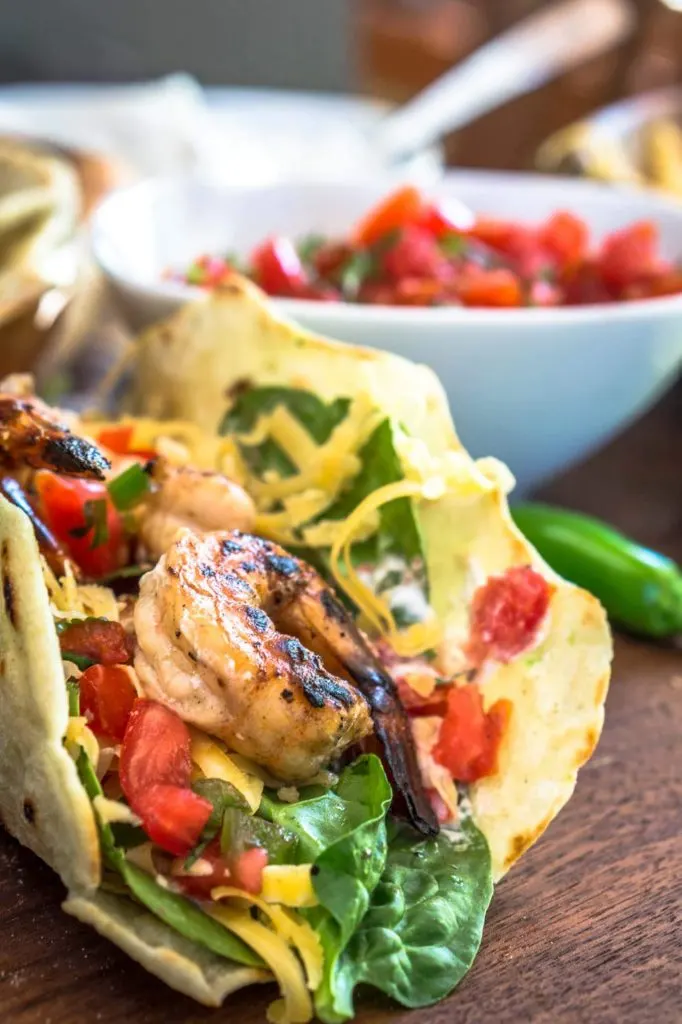 Taco shell filled with grilled shrimp, tomatoes, cheese, and jalapeno. A white bowl of pico sits in the background.
