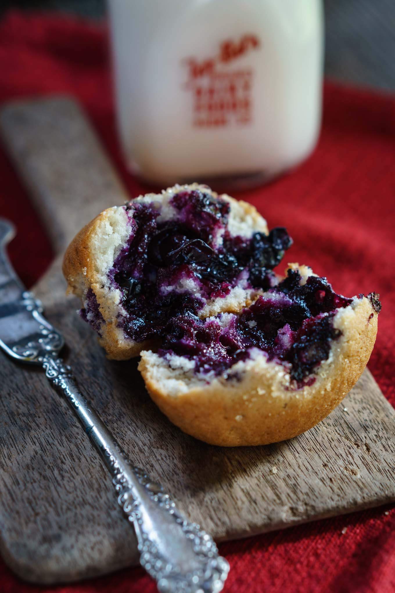 A muffin loaded with blueberries is broken open and sits on an antique wooden butter paddle next to an antique butter knife over a red napkin with a bottle of milk sitting in the back.