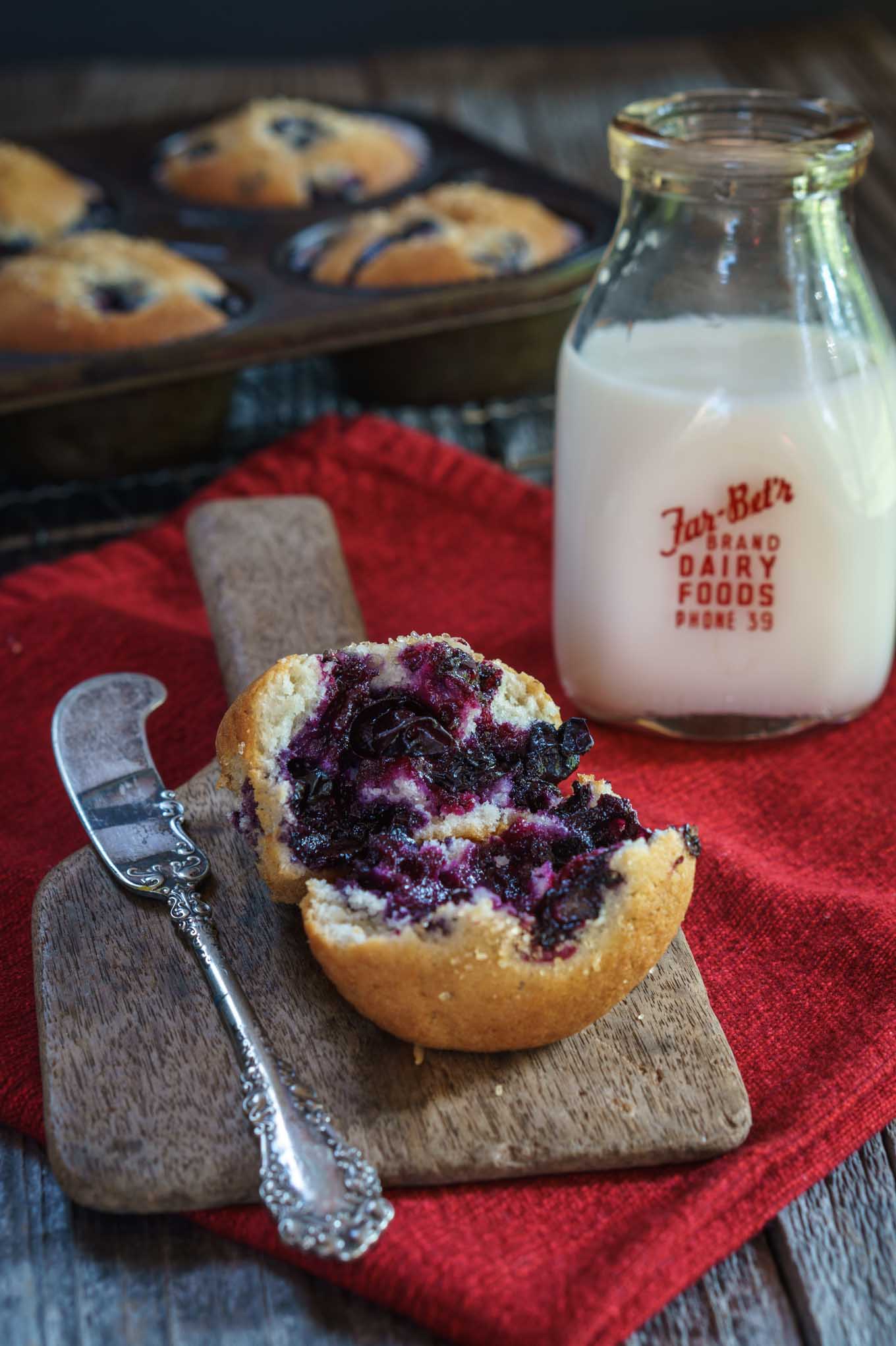 A broken open muffin loaded with blueberries sits on a wooden butter paddle nest to an antique butter knife. The paddle sits on a red napkin and a bottle of milk sits in the background.