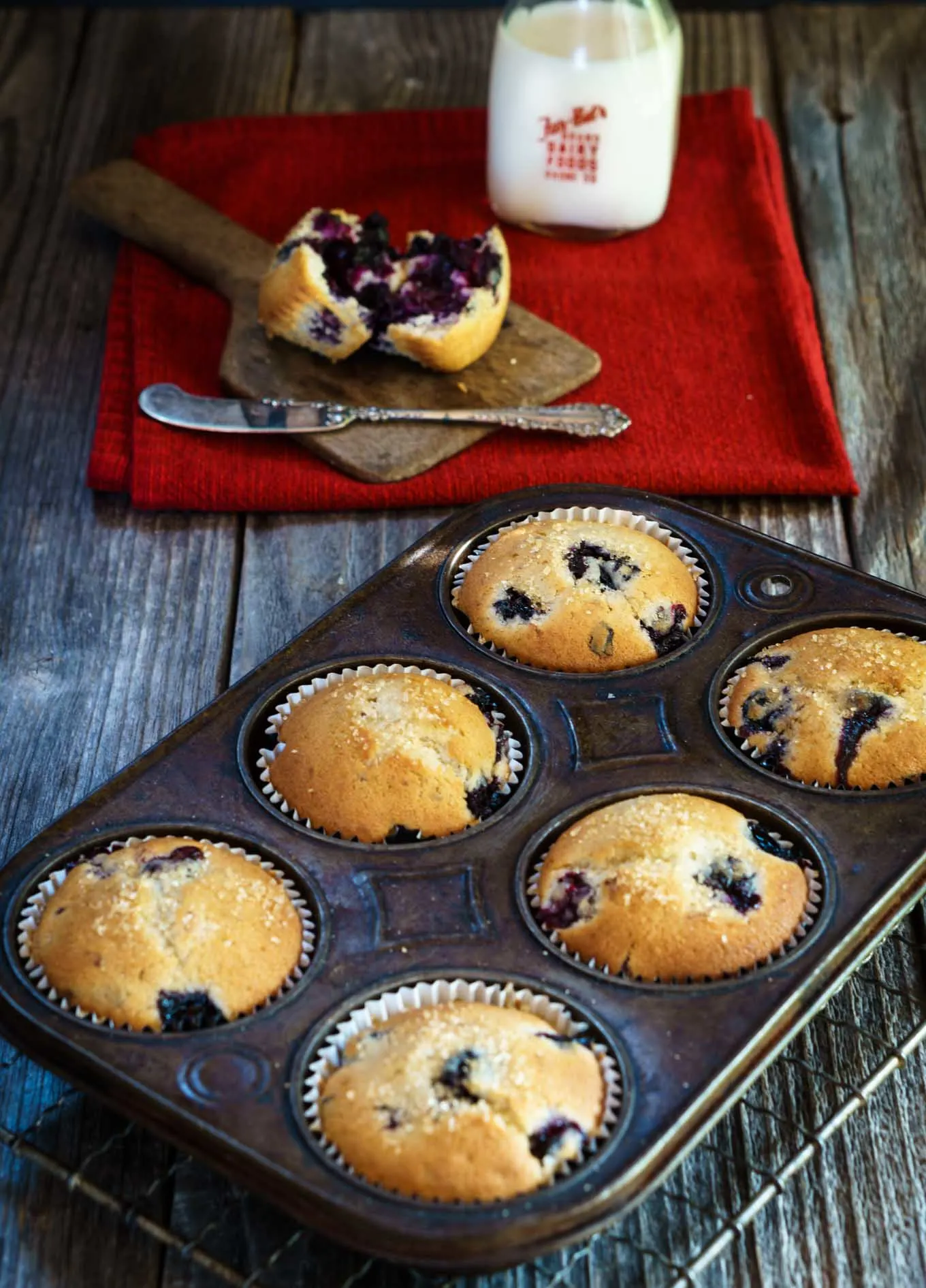 A muffin tin filled with baked blueberry muffins sitting on a cooling rack. A broken open muffin sitting on a wooden butter paddle and a bottle of milk sit in the background.