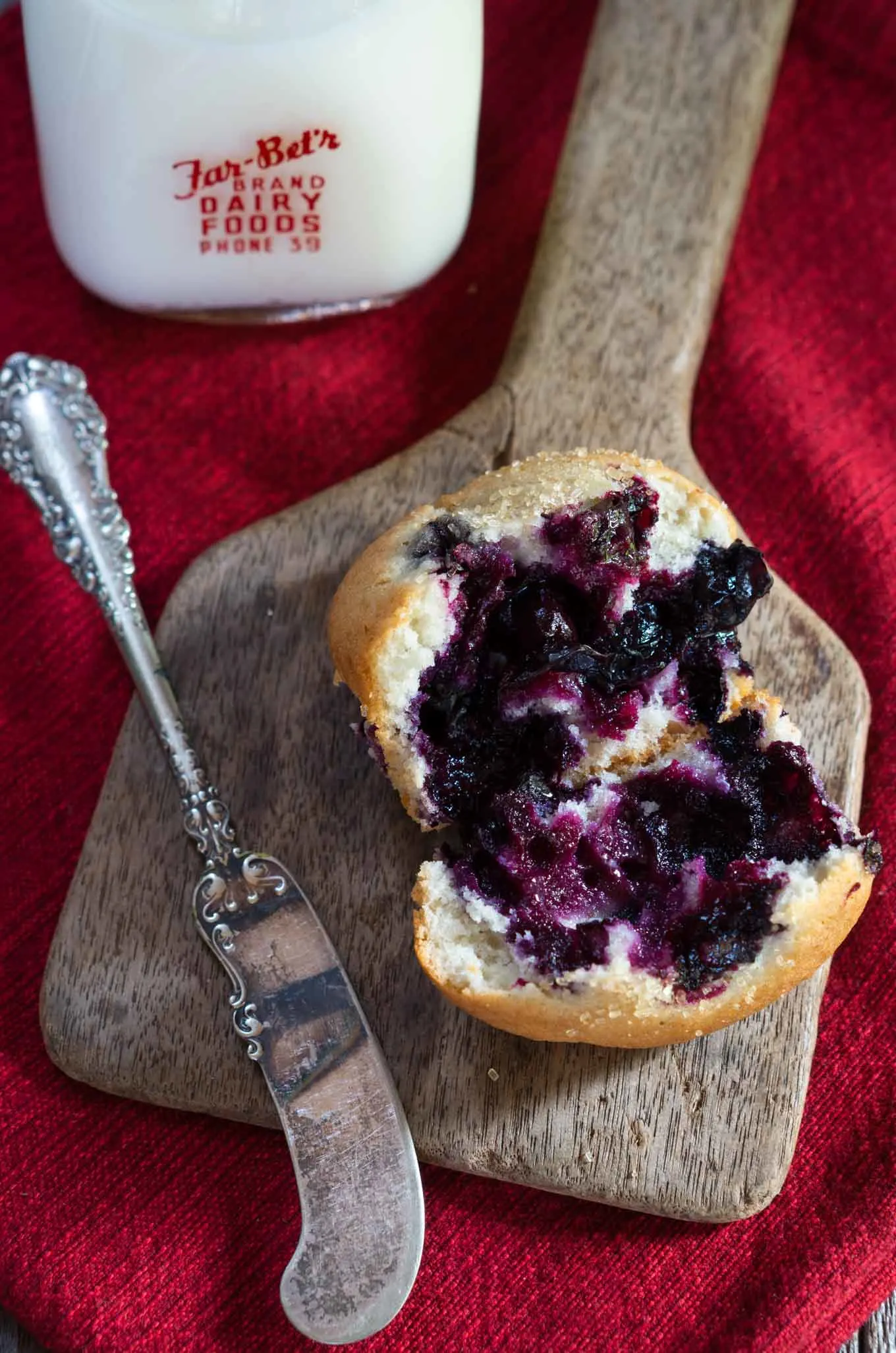 A broken open muffin loaded with blueberries sits on a wooden butter paddle nest to an antique butter knife. The paddle sits on a red napkin and a bottle of milk sits in the background.