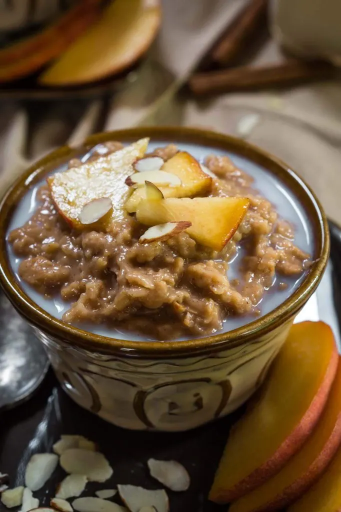 Side view of a bowl of Spiced Apple Steel Cut Oats sitting on a wooden cutting board. The bowl is sprinkled with nuts and raw apples. 