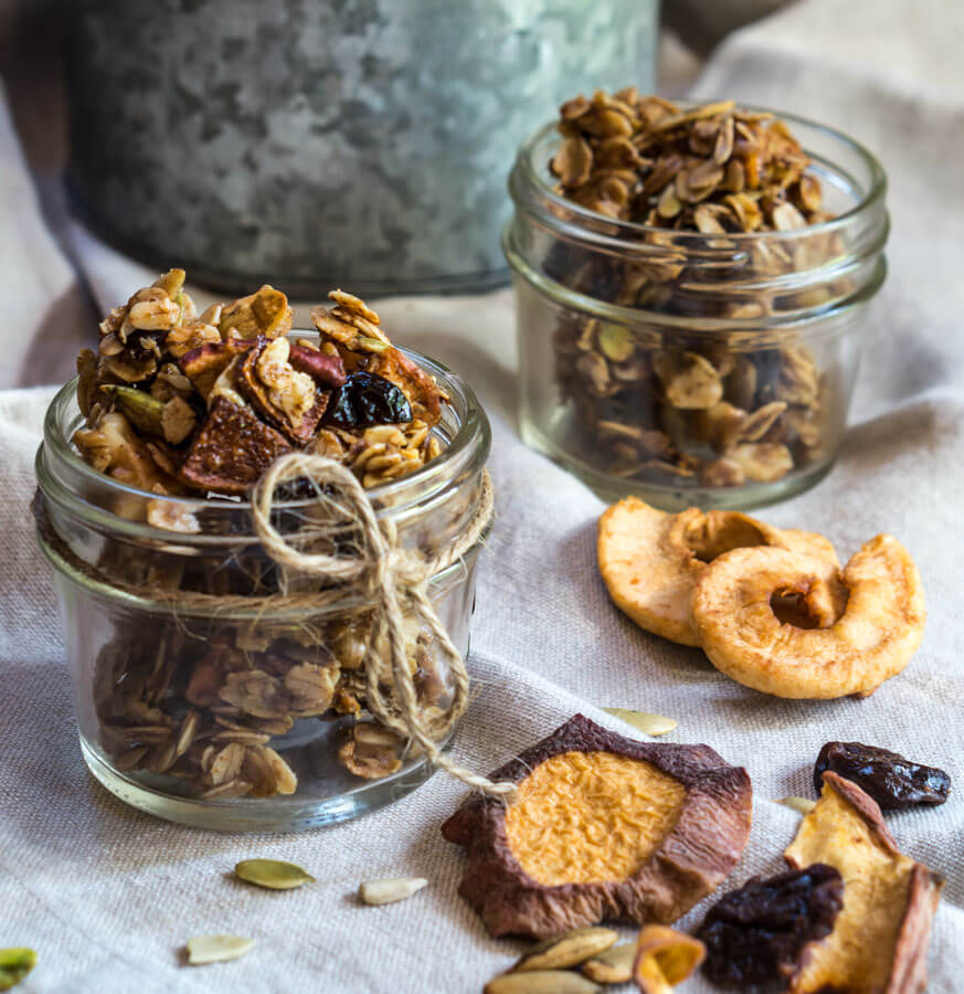 Two small jars of granola sit in small jars with a larger tin of granola in the background. Dried apples, cherries, and nuts sit in the foreground.