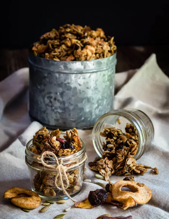 Two small jars of granola sit in small jars with a larger tin of granola in the background. Dried apples sit in the foreground with one sitting on its side.
