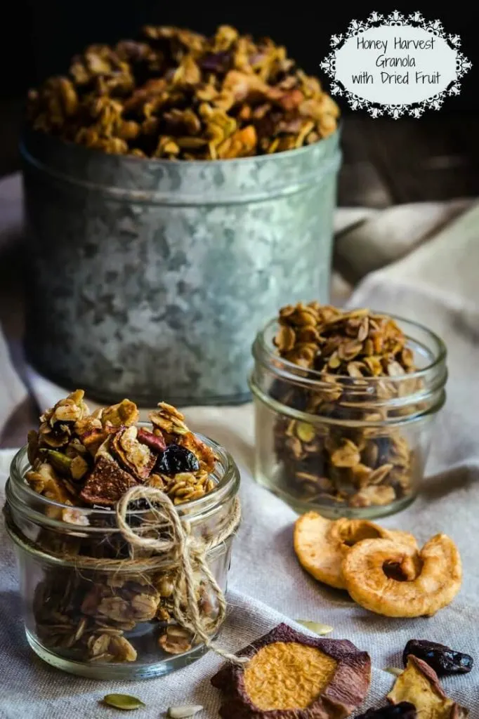 Two small jars of granola sit in small jars with a larger tin of granola in the background. Dried apples sit in the foreground.