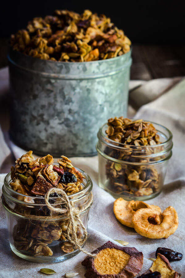Two small jars of granola sit in small jars with a larger tin of granola in the background. Dried apples sit in the foreground.