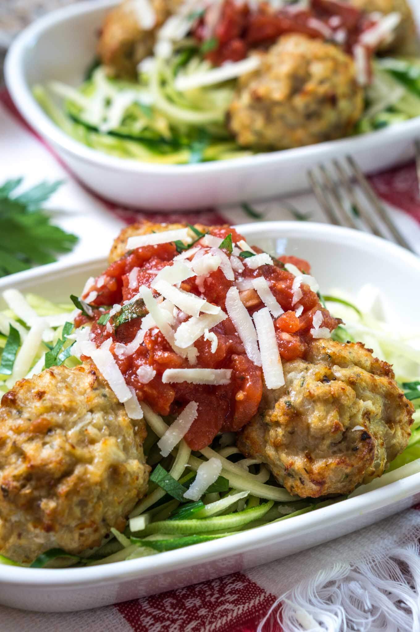 A white bowl of spiralized zucchini topped with chicken meatballs and marinara and garnished with parmesan. A second dish sits in the background.