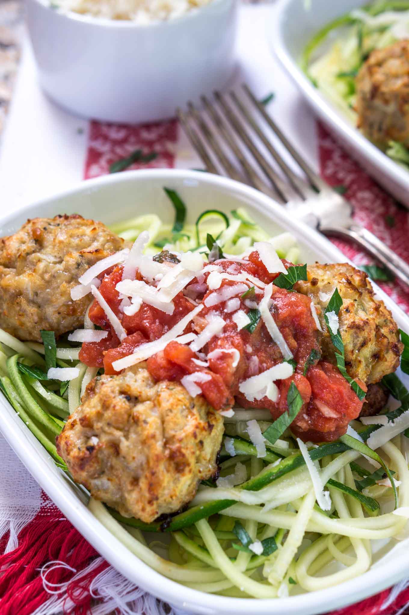  A white bowl of spiralized zucchini topped with chicken meatballs and marinara. A second dish and forks sit in the background.