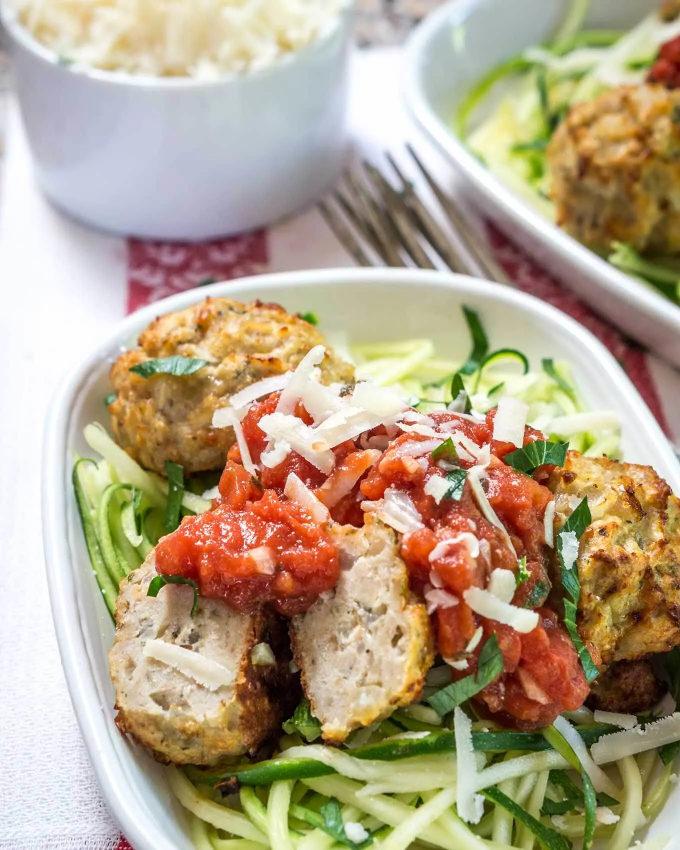 Top view of a white bowl of spiralized zucchini topped with chicken meatballs and marinara. Forks and another dish sit in the background.