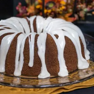 Bundt cake with icing drizzled over the top and dripping onto the serving platter with fall flowers sitting behind it.