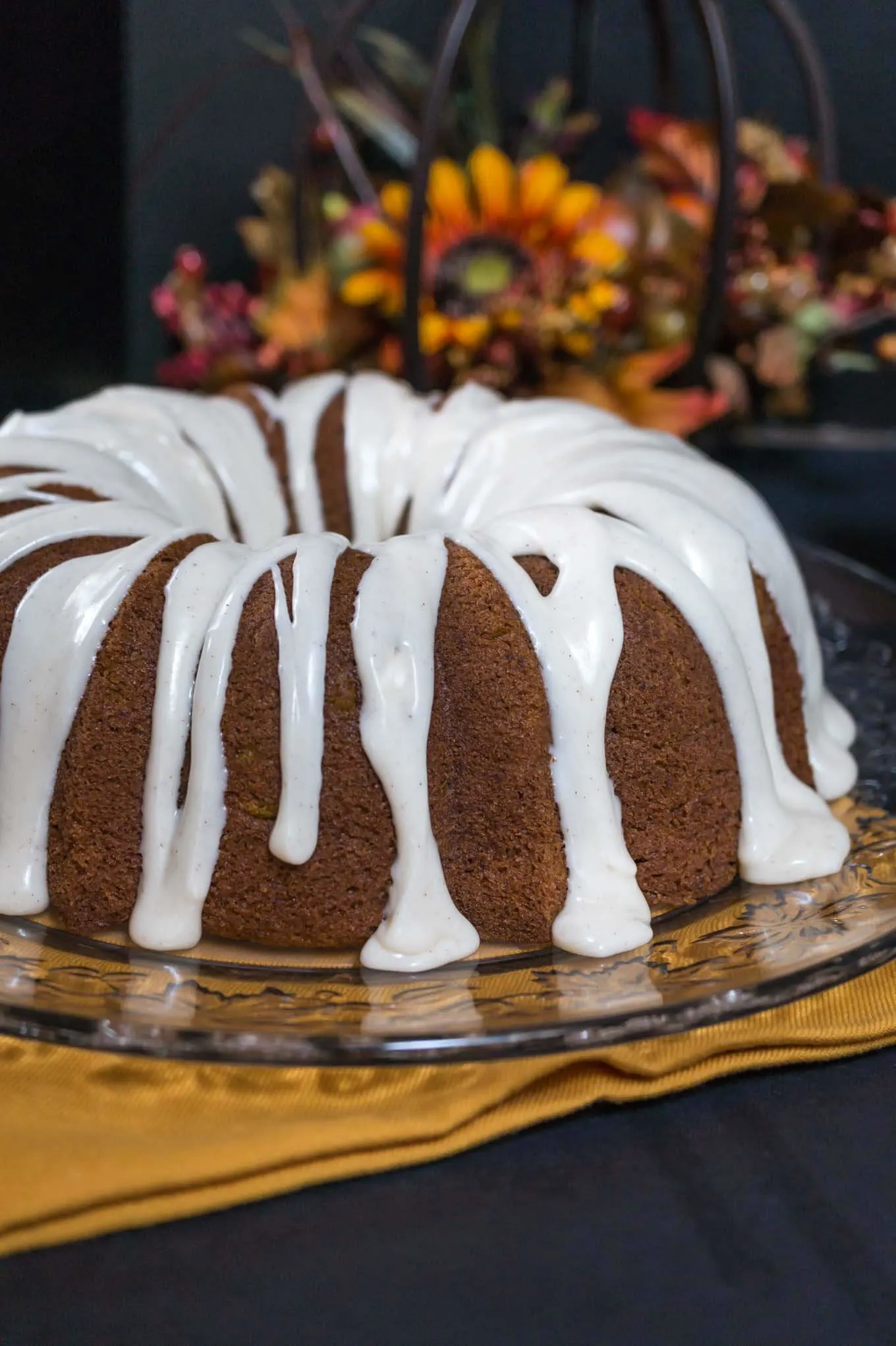 Pumpkin Bundt cake with icing drizzled over the top and dripping onto the serving platter with fall flowers sitting behind it.