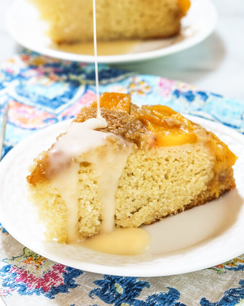 A piece of Upside-down Peach cake with a thin drizzle coming down from the top of the photo. The plate sits on a pastel napkin in front of a second plate.