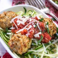 A white bowl of spiralized zucchini topped with chicken meatballs and marinara. A second dish and forks sit in the background.