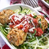 A white bowl of spiralized zucchini topped with chicken meatballs and marinara. A second dish and forks sit in the background.