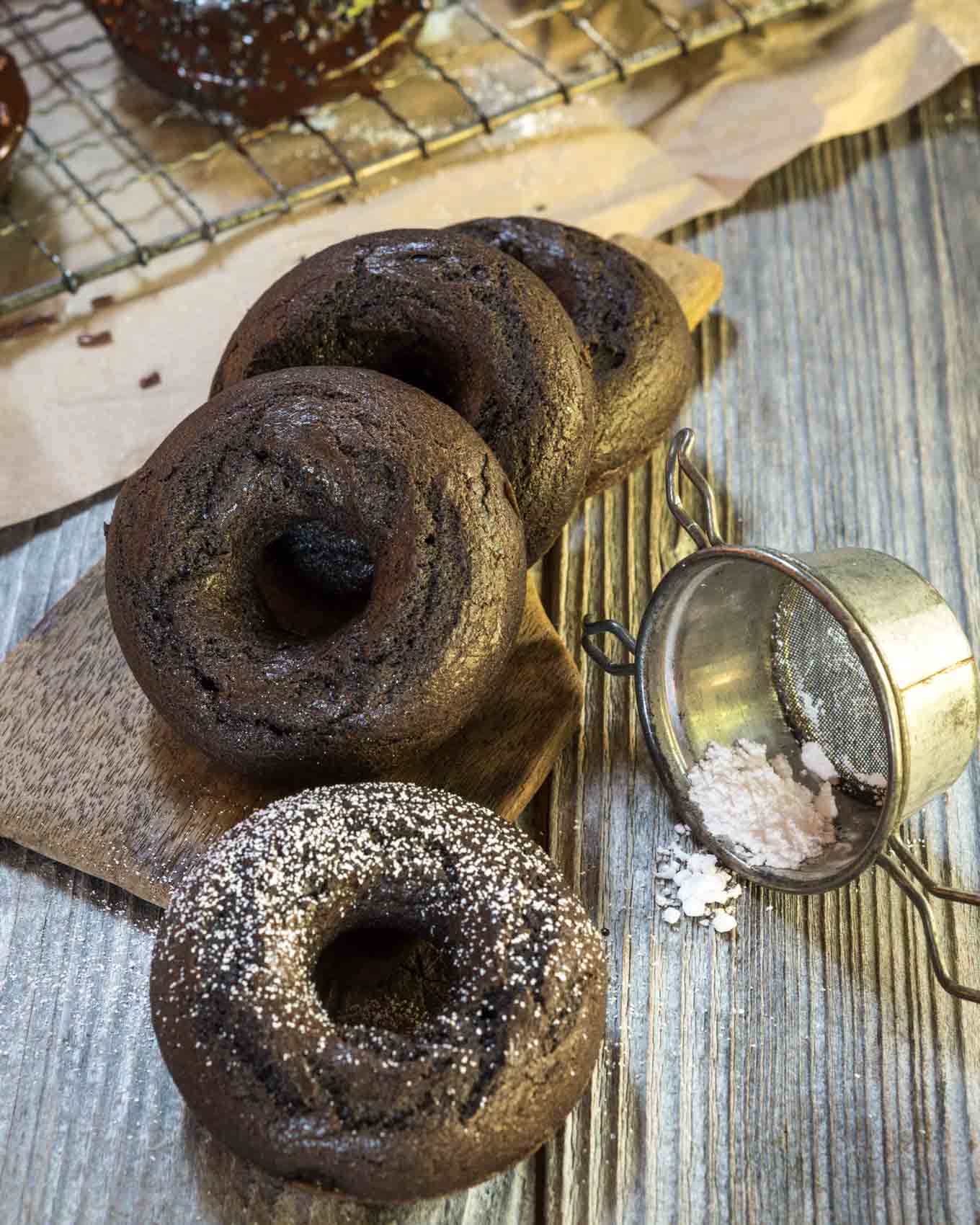 Thrde baked donuts leaning in a row. The front donut is sprinkled with powdered sugar next to an antique sifter.