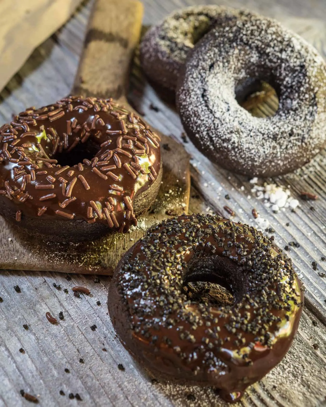 Three chocolate baked donuts. One dusted with powdered sugar, and two glazed with chocolate and sprinkled with chocolate sprinkles.