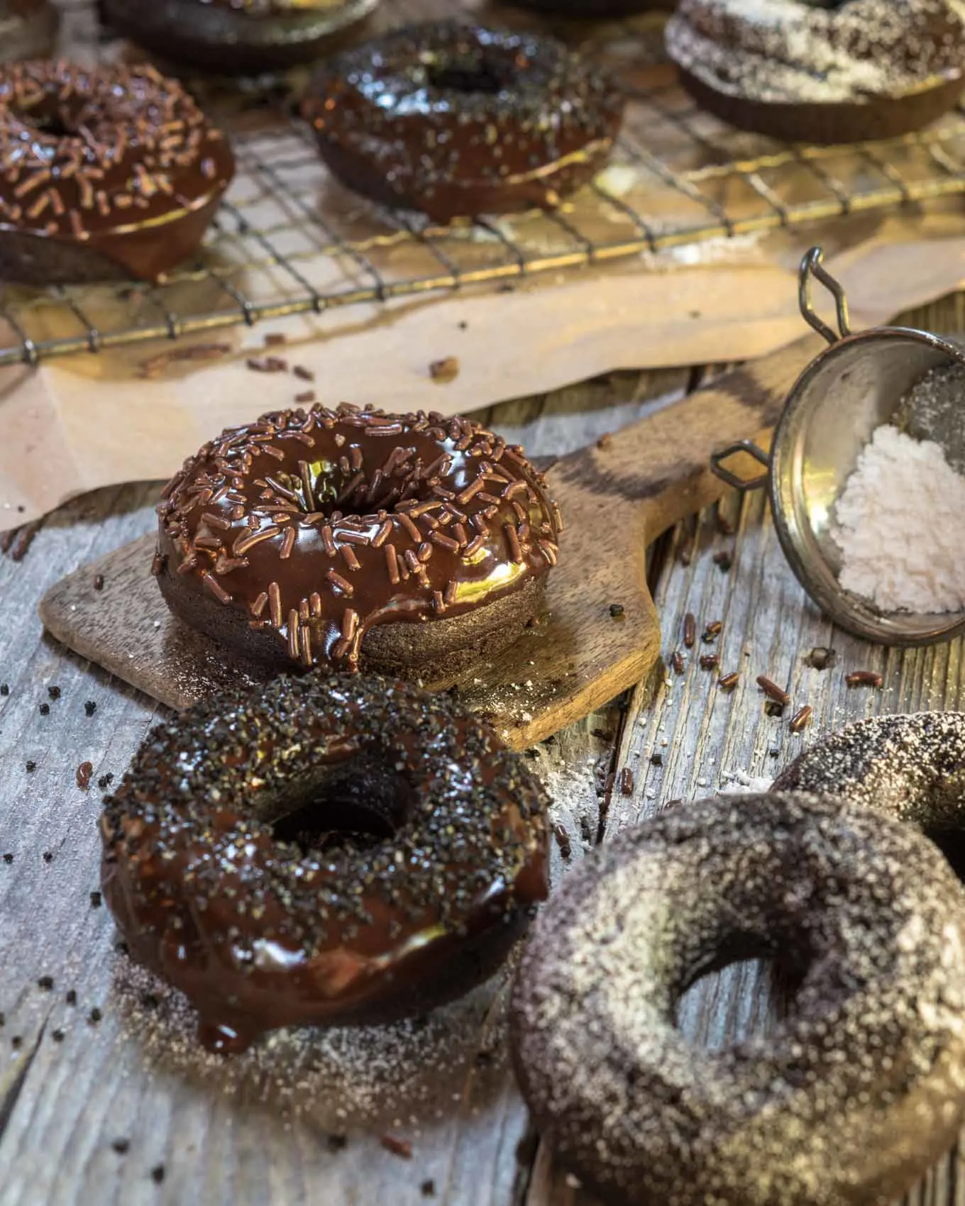 Baked chocolate donuts on a white wooden background that have been glazed and sprinkled or just dusted with powdered sugar. A cooling rack of more donuts sits in the background.