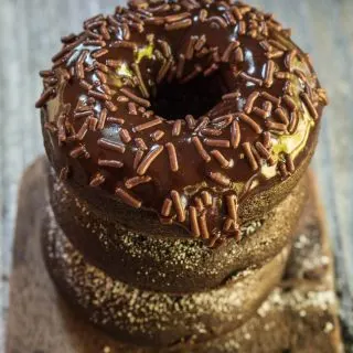 A stack of baked chocolate donuts sitting on an antique butter paddle. The top donut is glazed with chocolate and sprinkled with chocolate sprinkles.