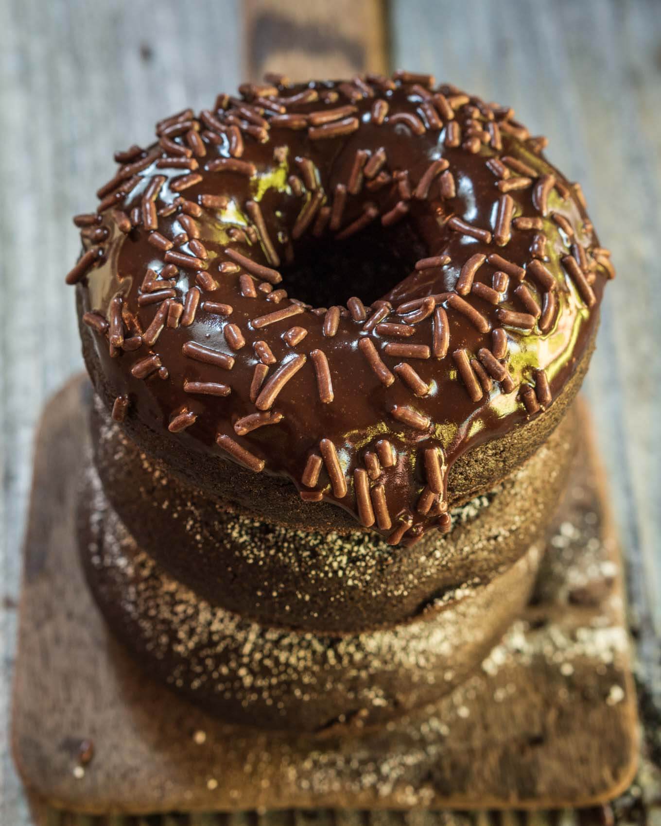 A stack of baked chocolate donuts sitting on an antique butter paddle. The top donut is glazed with chocolate and sprinkled with chocolate sprinkles.