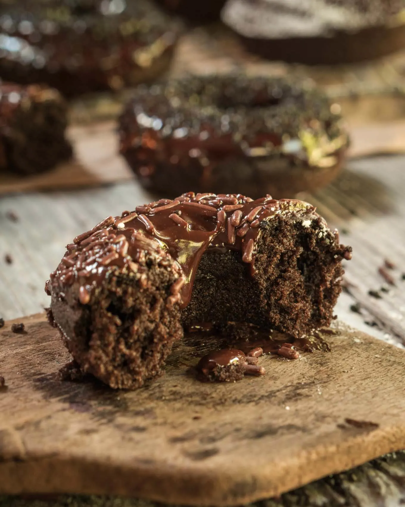 A side view of half of a baked chocolate donut sitting on a wooden butter paddle. A whole donut sits in the background.