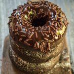 A stack of chocolate donuts sitting on an antique butter paddle. The top donut is dipped in chocolate and chocolate sprinkles.