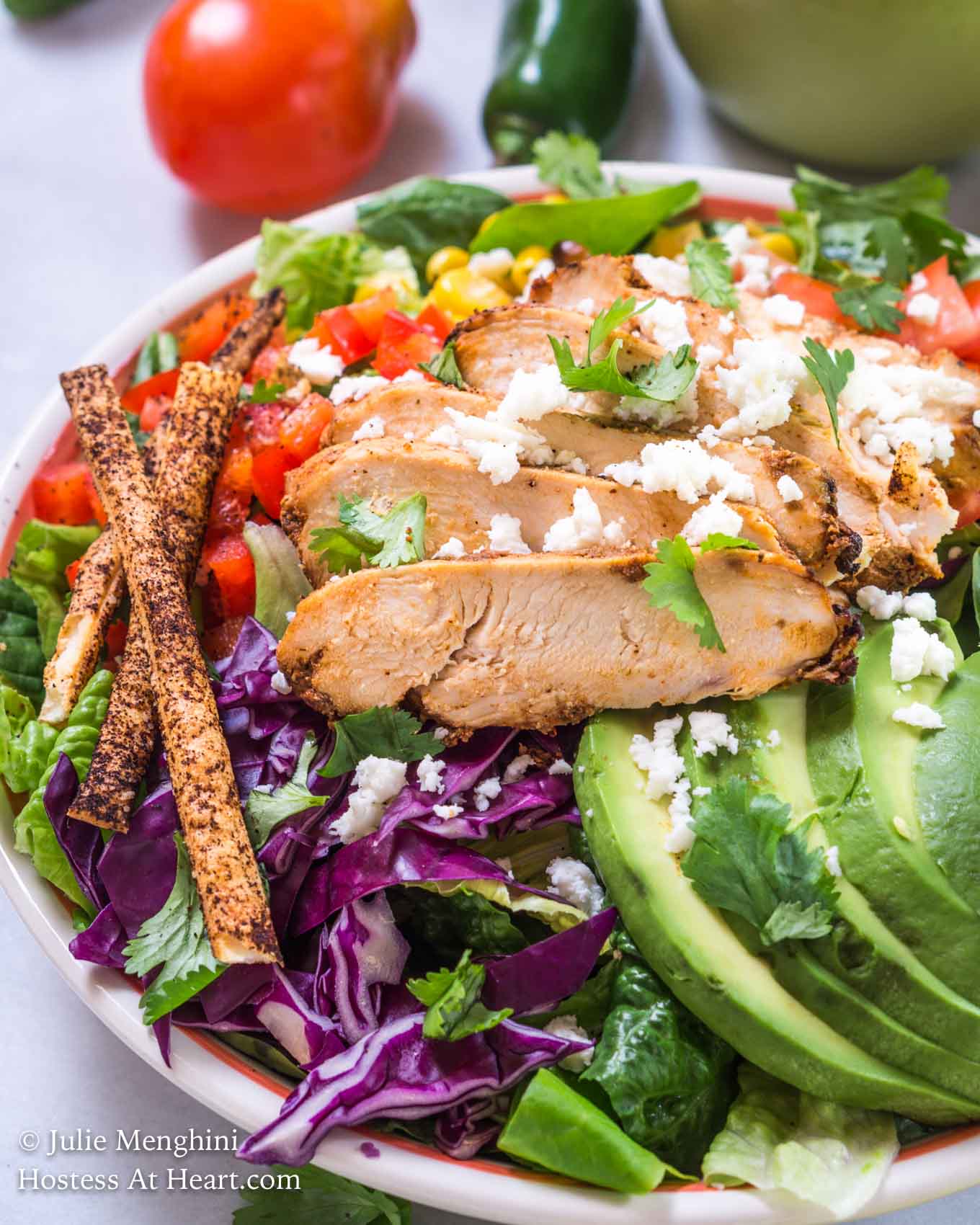 Angled view of a green salad topped with slices of chipotle grilled chicken, sliced avocado, shredded purple cabbage, diced tomato, grilled corn, and tortilla straws and then garnished with fresh cilantro. Fresh tomato and jalapenos sit in the background.