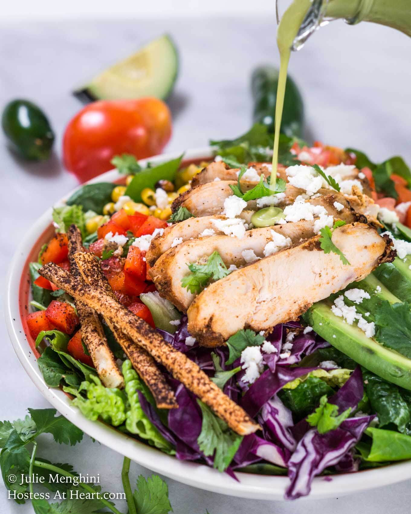 Side angle of a green salad topped with slices of chipotle grilled chicken, sliced avocado, shredded purple cabbage, diced tomato, grilled corn, and tortilla straws and then garnished with fresh cilantro being drizzled with dressing. A fresh tomato, jalapeno, and avocado sit in the background, 