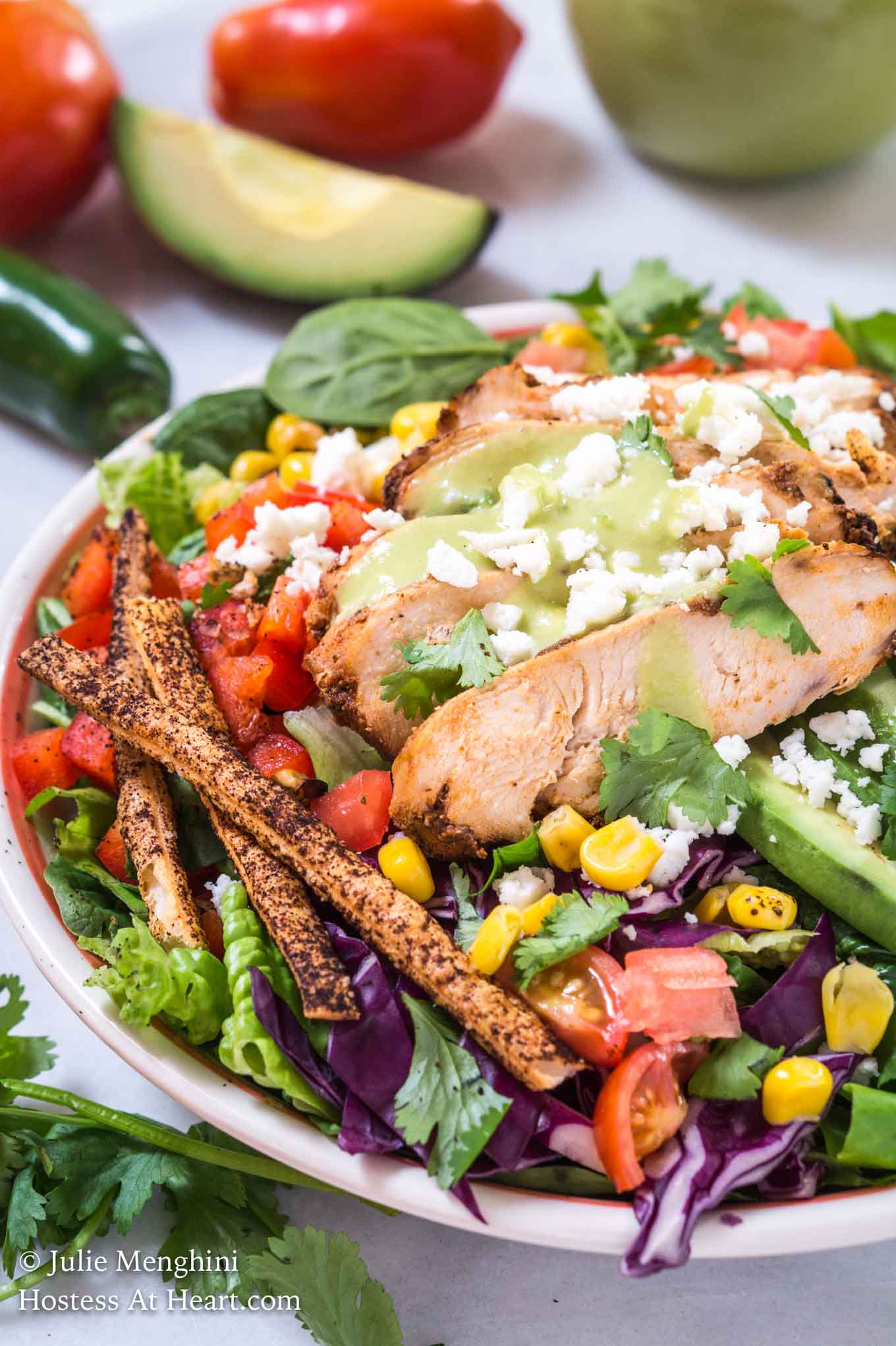 Partial front view of a green salad topped with slices of chipotle grilled chicken, sliced avocado, shredded purple cabbage, diced tomato, grilled corn, and tortilla straws and then garnished with fresh cilantro. A fresh tomato, jalapeno, and avocado sit in the background.