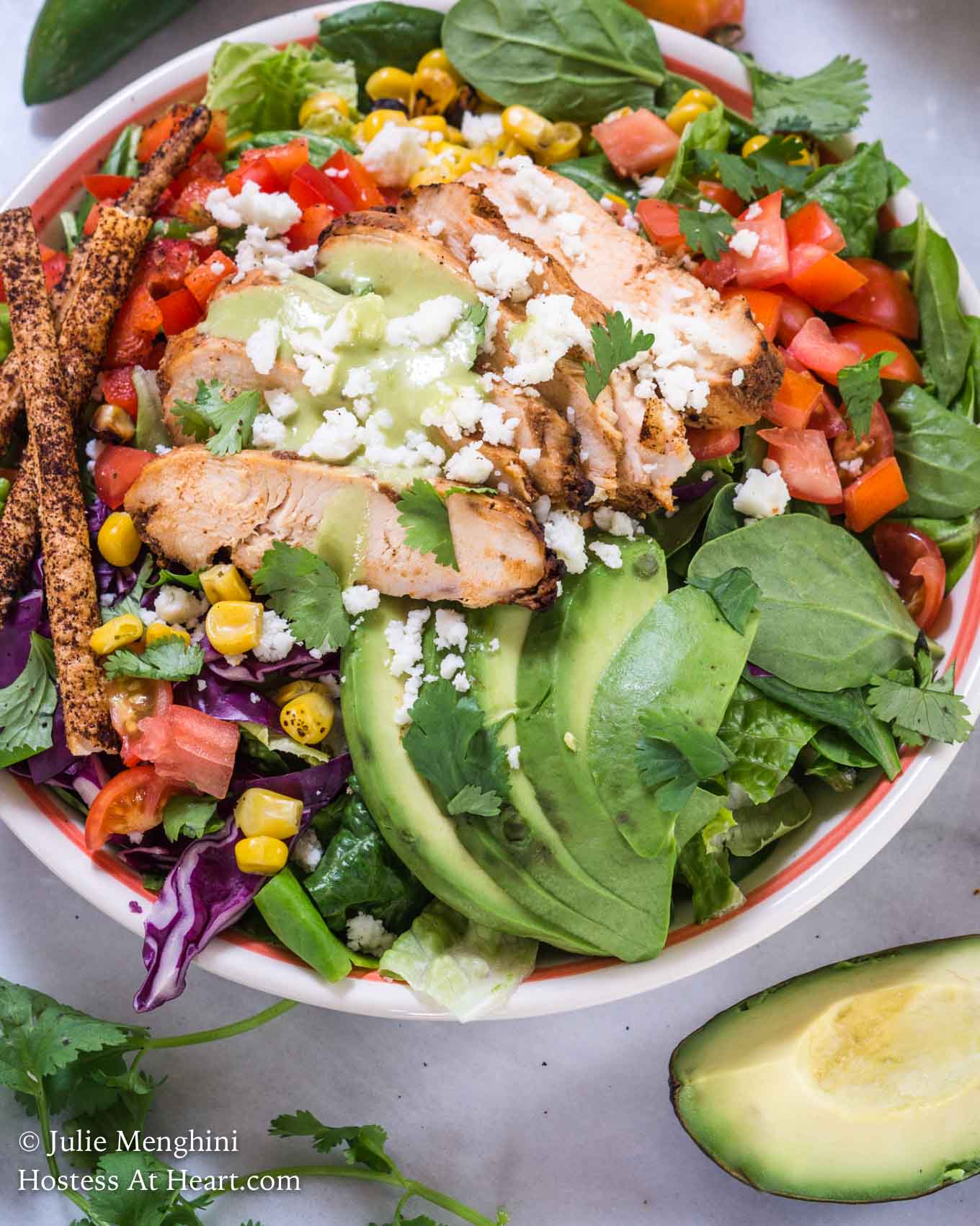 Top-down view of a green salad topped with slices of chipotle grilled chicken, sliced avocado, shredded purple cabbage, diced tomato, grilled corn, and tortilla straws and then garnished with fresh cilantro. A cut avocado sits in the foreground.