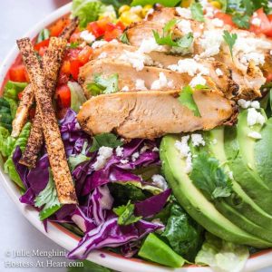 A salad topped with sliced chicken, avocados, diced red pepper, roasted corn, and tomatoes and garnished with cheese and tortilla straws.