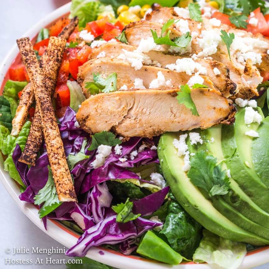 A salad topped with sliced chicken, avocados, diced red pepper, roasted corn, and tomatoes and garnished with cheese and tortilla straws.