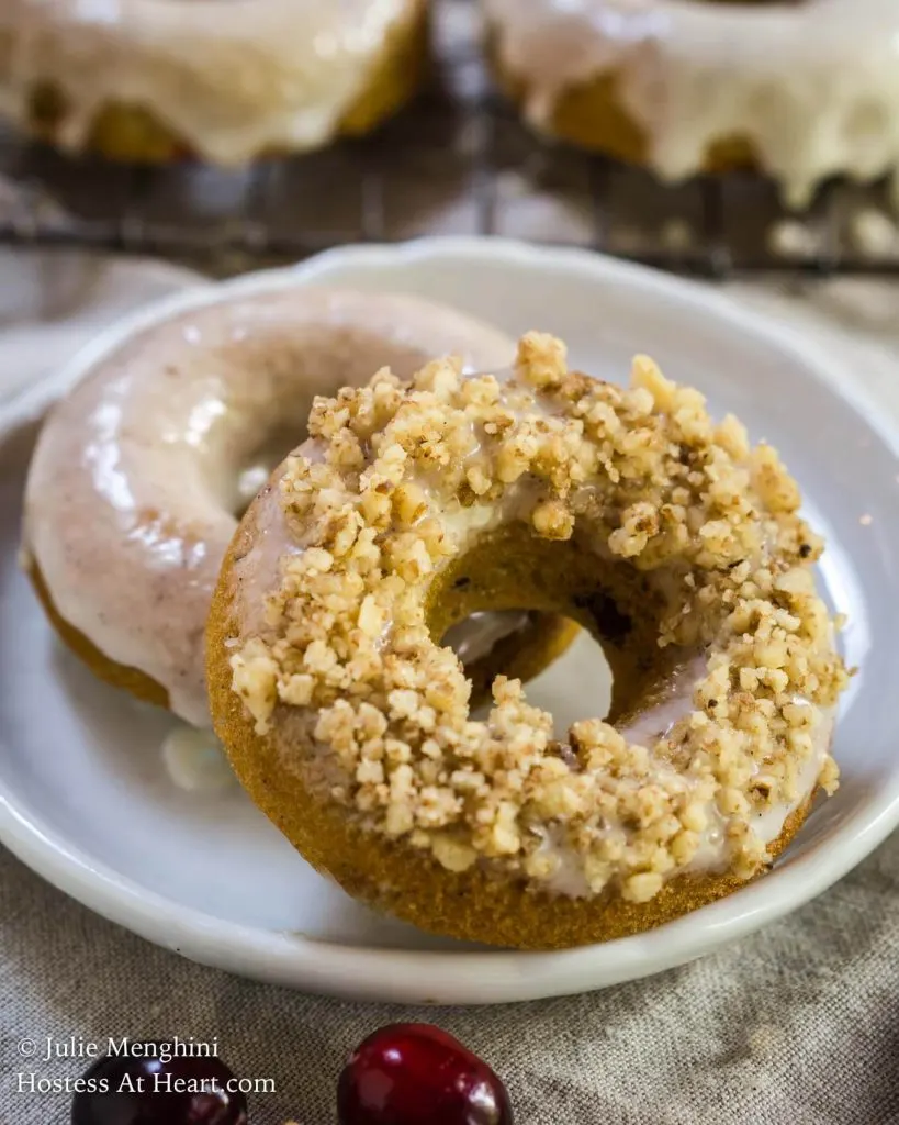 Two baked donuts on a white plate. The front forward one is topped with glaze and rolled in nuts. The back one is glazed. A cooling rack holding more donuts sit in the background.