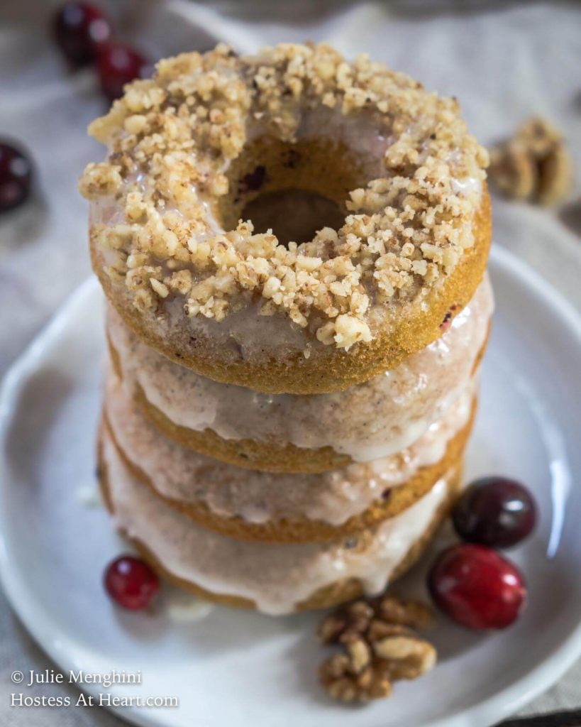 A stack of baked and glazed donuts tower over a white plate. Fresh cranberries and walnuts sit next to the donuts.