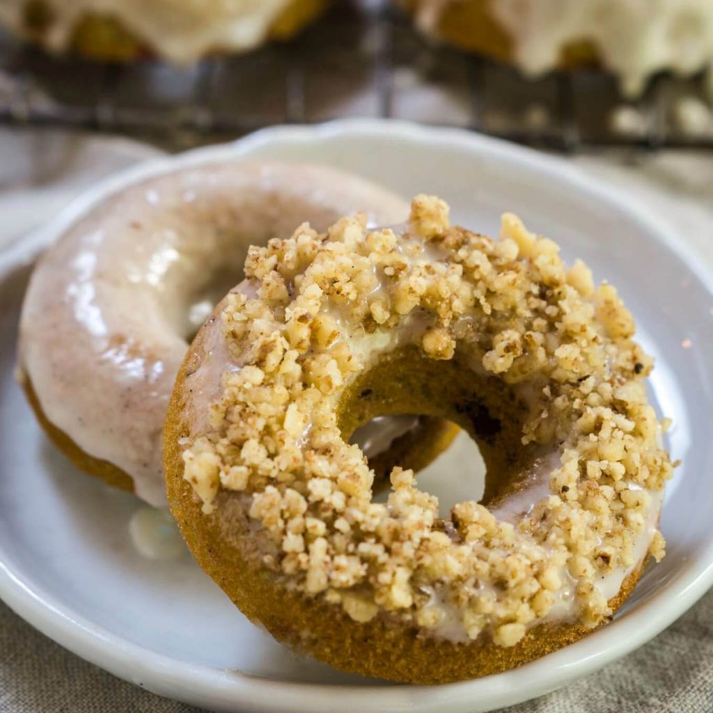 Two baked glazed donuts sit on a white plate. The front one is topped with ground walnuts. A cooling wrack with more baked and glazed donuts sit in the background.