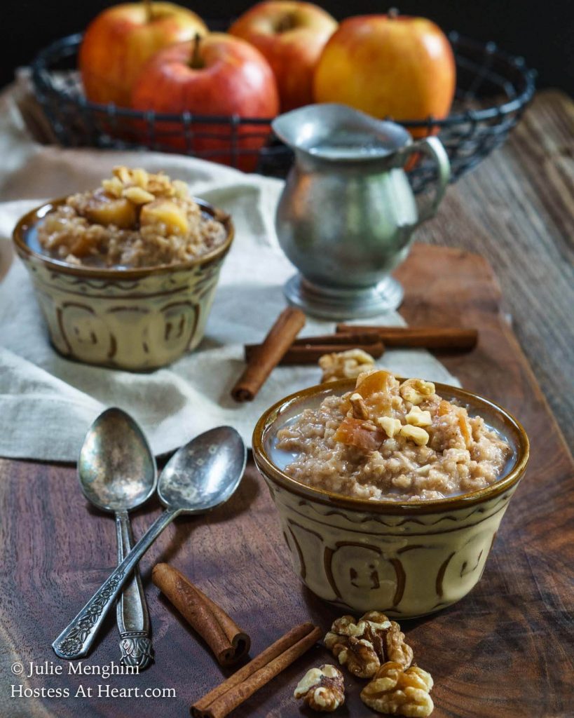 Side angle of two bowls of steel cut oats topped with cooked cinnamon apples and nuts. Two spoons sit next to the dishes and a milk pitcher sits in the back.