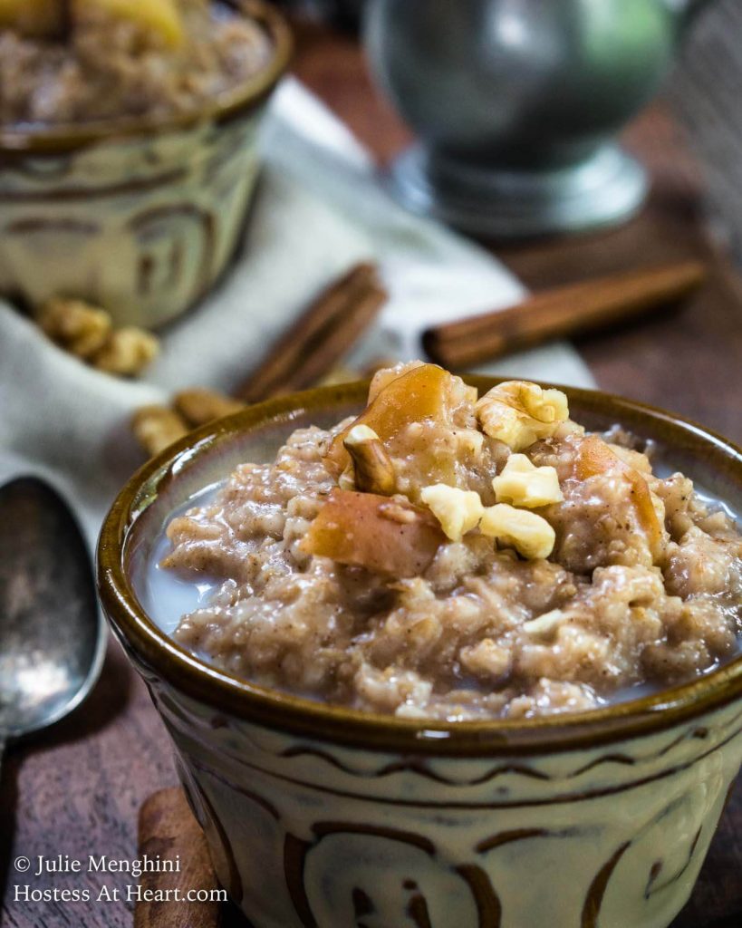 Side view of a bowl of Spiced Apple Steel Cut Oats sitting on a wooden cutting board. The bowl is sprinkled with nuts and raw apples. A spoon sits next to the bowl and another bowl sits in the background.