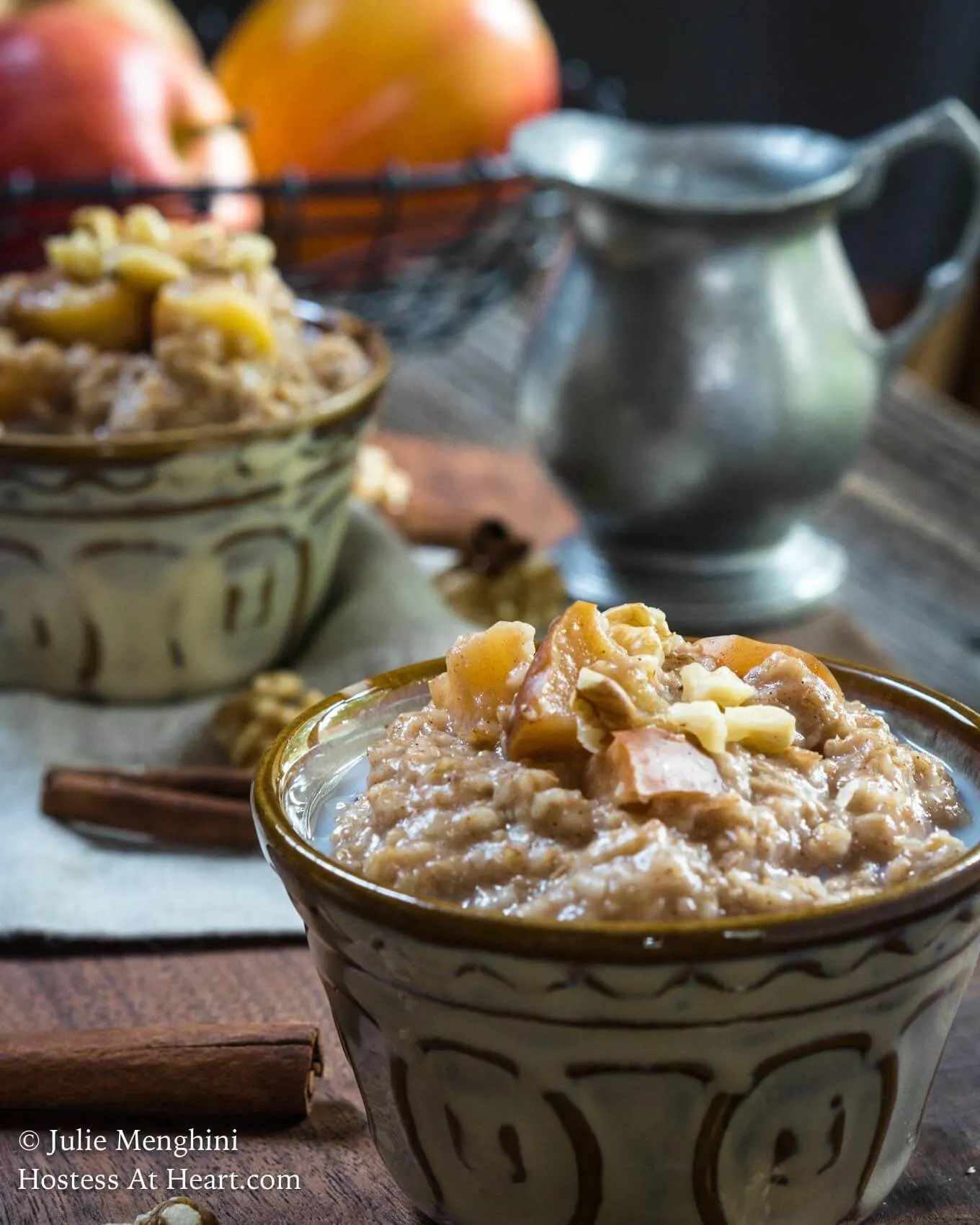 Side view of a bowl of Spiced Apple Steel Cut Oats sitting on a wooden cutting board. The bowl is sprinkled with nuts and raw apples. A spoon sits next to the bowl and another bowl and a pitcher of milk sits in the background.