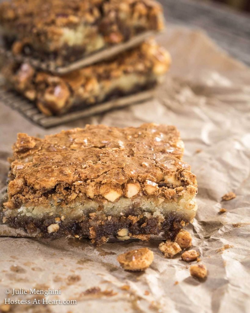 A Hazelnut Cookie Bar sitting on a piece of parchment paper with fresh hazelnuts scattered in the foreground. A stack of the bars sits in the background.