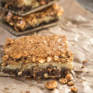 A bar of a Hazelnut Cookie bar sits on a piece of parchment paper with a sprinkling of hazelnuts in front of it and a stack of bars behind it.