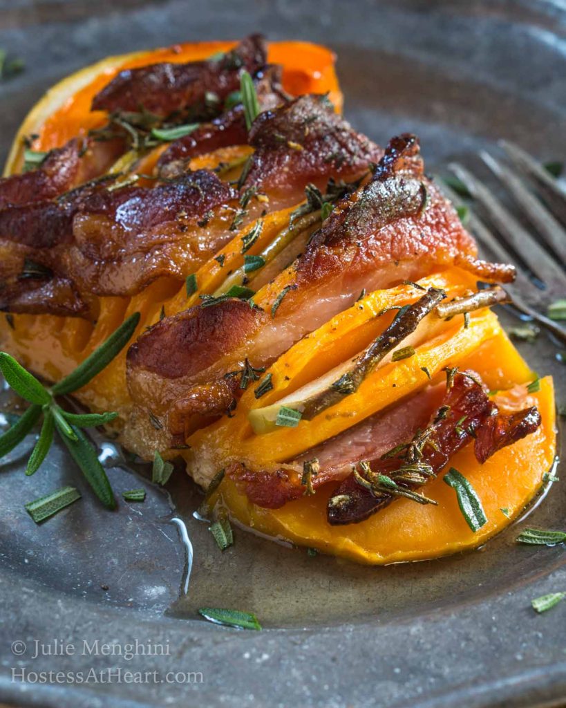 A metal plate holding a butternut squash that is sliced and filled with bacon and apple then sprinkled with rosemary. A fork sits off to the side.