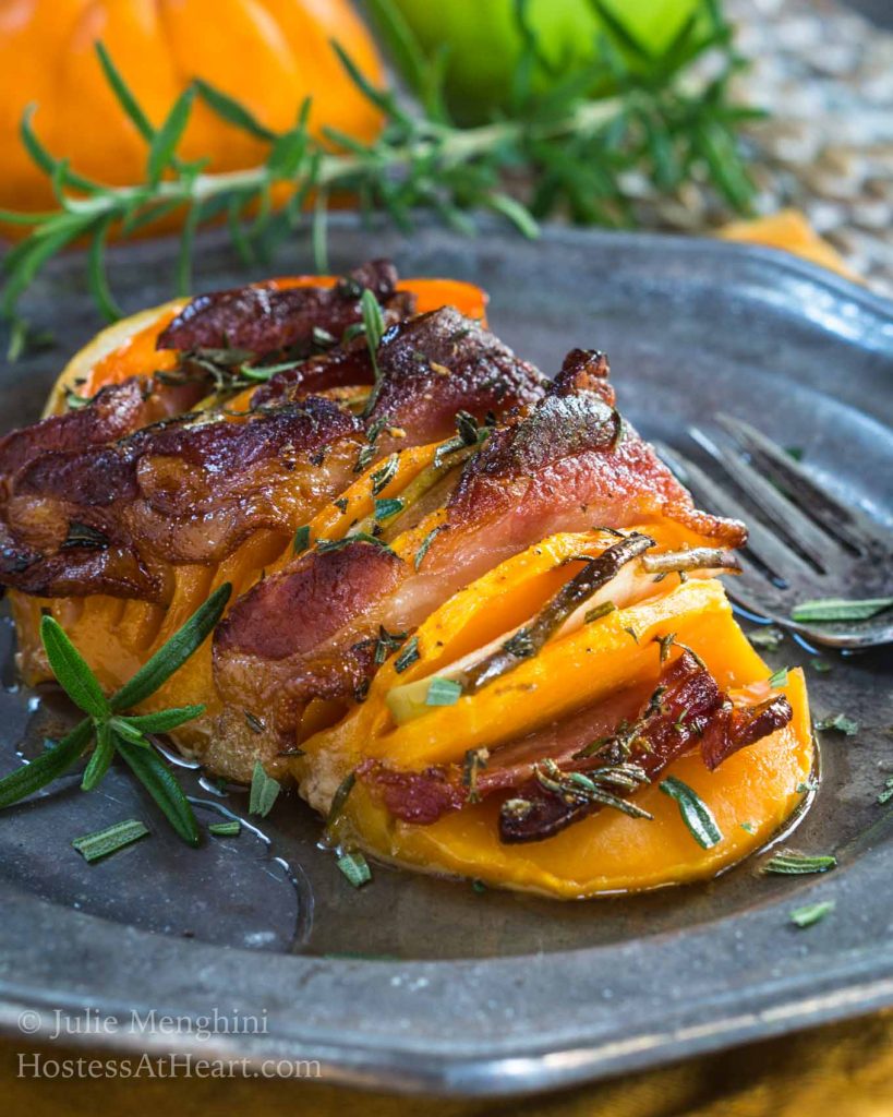 A metal plate holding a butternut squash that is sliced and filled with bacon and apple then sprinkled with rosemary. A fork sits off to the side.