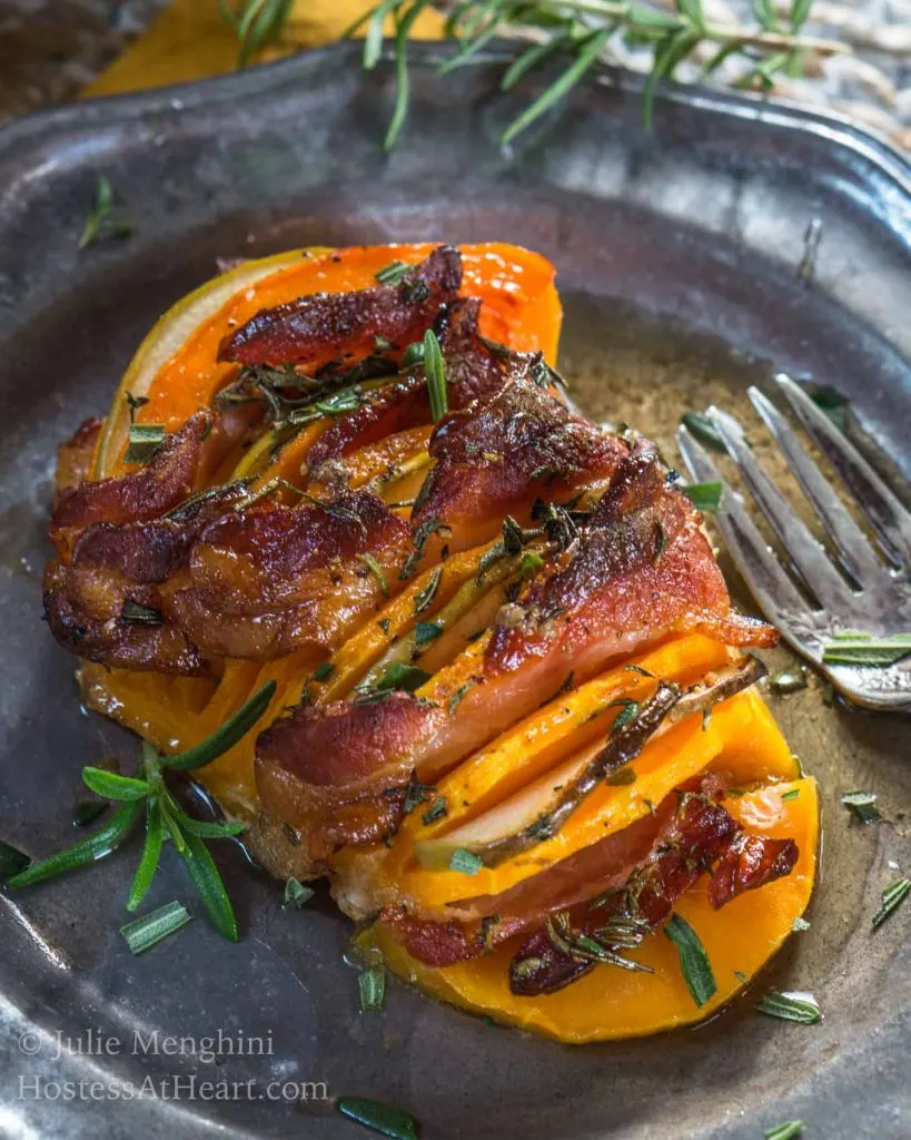 A metal plate holding a butternut squash that is sliced and filled with bacon and apple then sprinkled with rosemary. A fork sits off to the side.