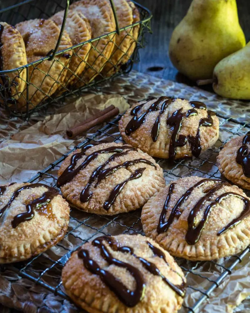 A cooling rack over a wooden matt filled with Pear Hand Pies drizzled with dark chocolate. Two raw pears and a wire basket of pies sit in the background.