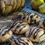  Round-shaped pear hand pies that have been drizzled with dark chocolate over a cooling rack. A basket of additional pies sits in the back next to fresh pears.