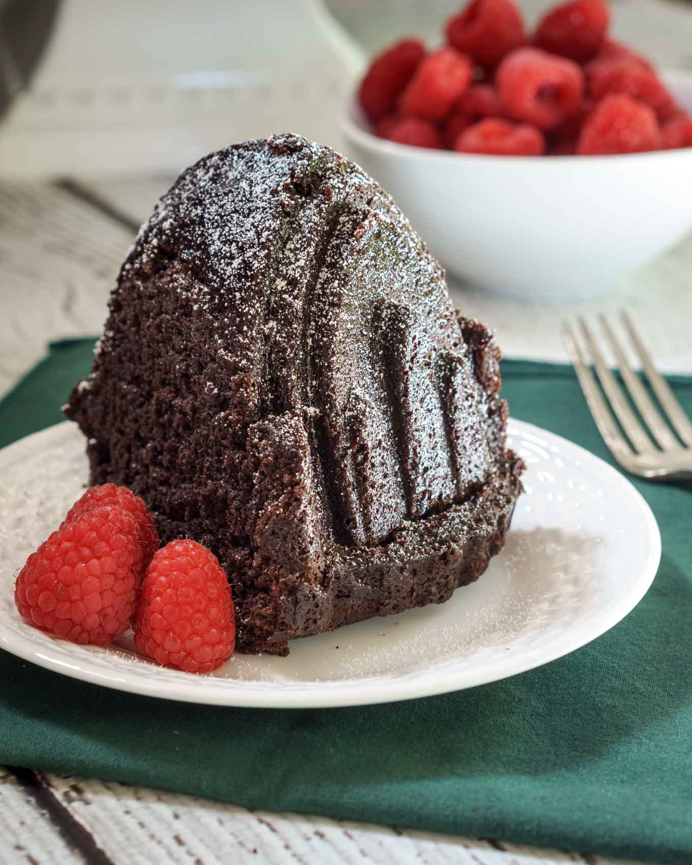 A slice of a raspberry glazed chocolate raspberry cake sitting on a white plate. Fresh raspberries sit on a plate and in a bowl behind the slice.