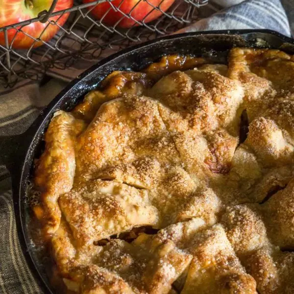 op view of 3/4 of an apple pie baked in a cast-iron skillet with a basket of fresh apples sitting behind it 