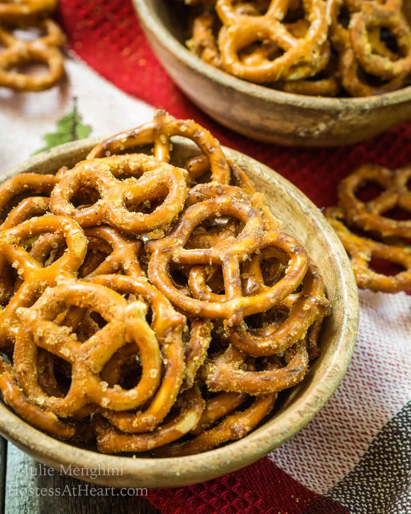 Top-down view of a wooden bowl filled with pretzels that have been baked in a spicy Nacho Cheese seasoning. A second bowl of pretzels sits in the background over a red checked towel.