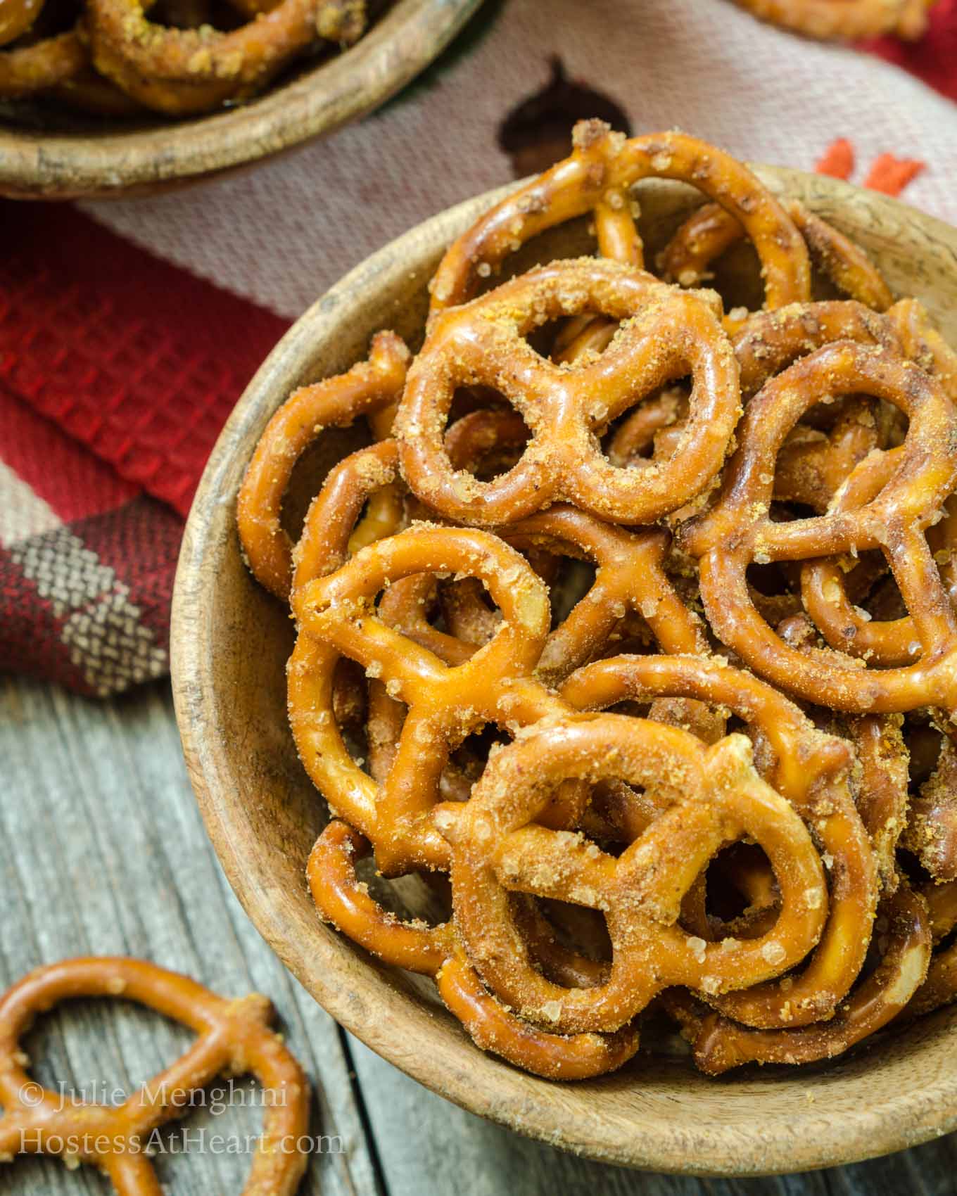 Top-down view of a wooden bowl filled with pretzels that have been baked in a spicy Nacho Cheese seasoning. A second bowl of pretzels sits in the background over a wooden background.
