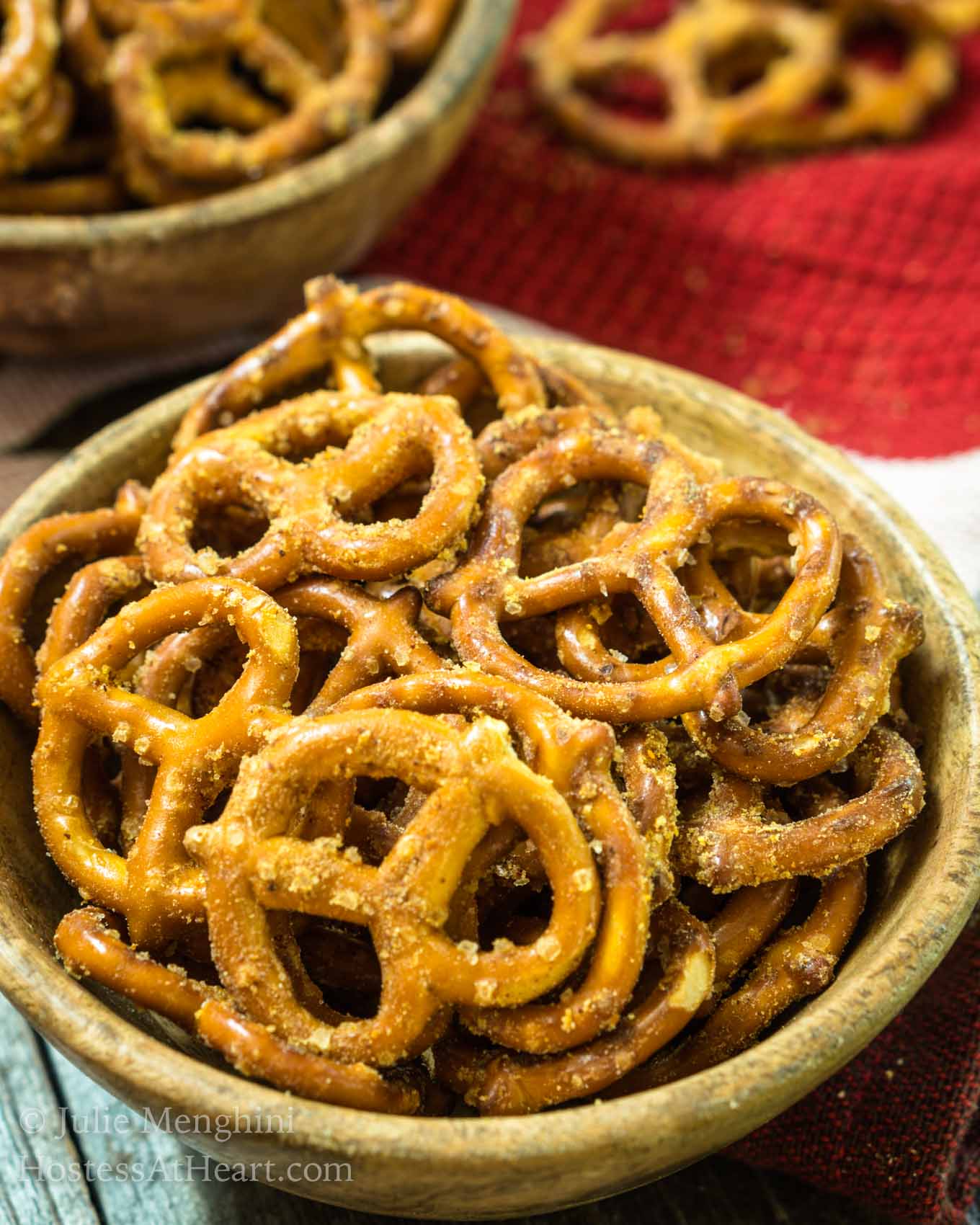 Angled view of a wooden bowl filled with pretzels that have been baked in a spicy Nacho Cheese seasoning. A second bowl of pretzels sits in the background over a wooden background.