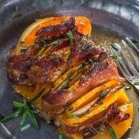 A metal plate holding a butternut squash that is sliced and filled with bacon and apple then sprinkled with rosemary. A fork sits off to the side.